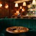 Intérieur élégant d’un club parisien en soirée, avec fauteuils en velours vert, lumière ambrée, boiseries sombres et portefeuille posé près de quelques pièces.