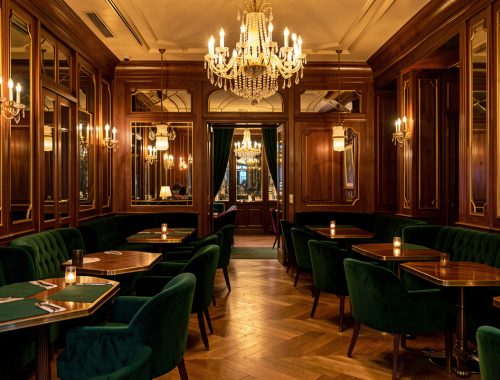 Salon parisien élégant aux tables en velours vert, fauteuils raffinés, boiseries et lustres dorés dans une ambiance chaleureuse de week-end.