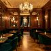 Salon parisien élégant aux tables en velours vert, fauteuils raffinés, boiseries et lustres dorés dans une ambiance chaleureuse de week-end.